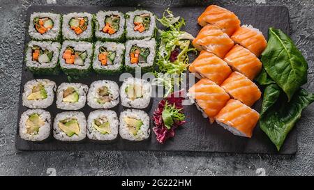 Lebensmittelbanner: Satz von verschiedenen Rollen auf einem Schneidebrett aus schwarzem Stein. Brötchen mit Lachs, Gemüse Sushi Maki-Brötchen und frischem Salat. Stockfoto