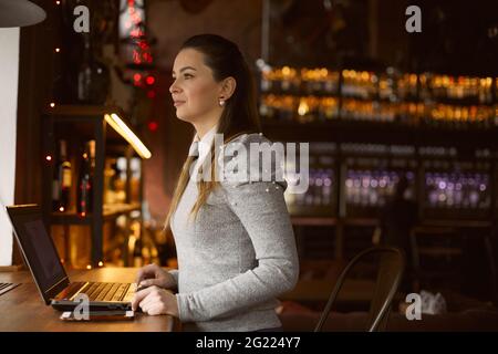 Das junge Mädchen schaut aus dem Fenster, arbeitet in einem Café hinter einem Laptop. Freiberufliches Konzept. Stockfoto