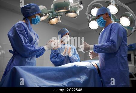 Ärzte stehen nach der Operation beim Patienten. Chirurg tut Daumen nach oben Geste, während weibliche Assistenten Blick auf den Mann und applaudieren. Konzept der Medizin, plastische Chirurgie, erfolgreiche Teamarbeit. Stockfoto