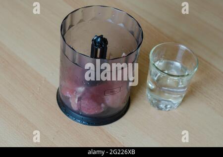 Schritt für Schritt Zubereitung von hausgemachtem gehacktem Schinken. Schritt 4: Ein Mixer mit Fleischstücken und einem Glas Frischwasser auf dem Tisch. Stockfoto