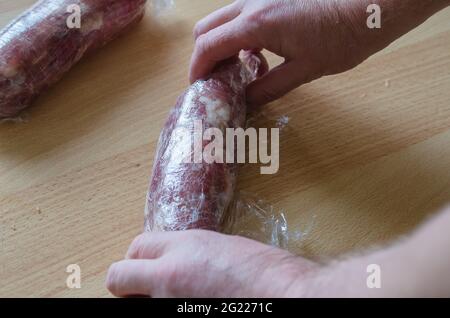 Schritt für Schritt Zubereitung von hausgemachtem gehacktem Schinken. Schritt 10: Die Hände eines Mannes drehen eine Portion mariniertes Fleisch in eine Rolle. Roher Schinken in Lebensmittelverpackung. C Stockfoto