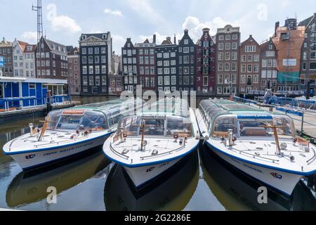 Amsterdam, Niederlande - 19. Mai 2021: Blick auf die ikonischen Häuser am Ufer des Damrak in der Innenstadt von Amsterdam mit Stadtrundfahrt Touristenboote in Stockfoto