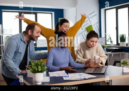 Verschiedene Kollegen feiern den Erfolg des Projekts und betrachten das Anheben der Hände auf dem Laptop. Multiethnisches Geschäftsteam mit Laptop und Papieren begeistert vom Projekt. Stockfoto