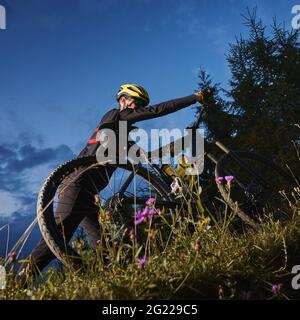 Low-Angle-Ansicht des Radfahrers, der sein Fahrrad bergauf schiebt. Hinterrad in Wildblumen im Vordergrund, grüne Fichten und Abendhimmel im Hintergrund. Konzept des Extremfahrens Stockfoto