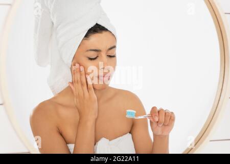 Junge afroamerikanische Frau mit Zahnbürste und Zahnschmerzen in der Nähe des Spiegels im Badezimmer