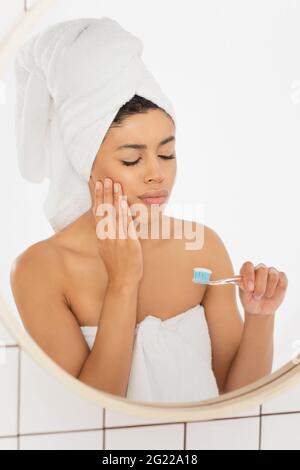 Junge afroamerikanische Frau mit Zahnbürste und Zahnschmerzen mit geschlossenen Augen in der Nähe des Spiegels im Badezimmer