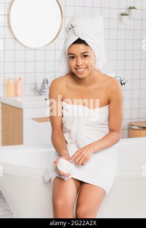 Lächelnde junge afroamerikanische Frau, die auf der Badewanne sitzt und im Badezimmer ein Glas mit Creme hält Stockfoto