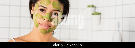 Junge Frau mit grüner Maske im Gesicht und Blick auf die Kamera im Badezimmer, Banner Stockfoto