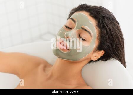 Entspannte junge afroamerikanische Frau mit Schlammmaske im Gesicht, die in der Badewanne liegt Stockfoto