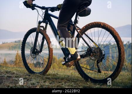 Horizontal geschnittene Momentaufnahme der Beine des Bikers, sitzend auf dem Fahrrad. Nahaufnahme auf dem Fahrrad, Räder im Schlamm, nach der morgendlichen Fahrt auf Bergwegen. Sportler, die den Moment des Sonnenaufgangs genießen Stockfoto