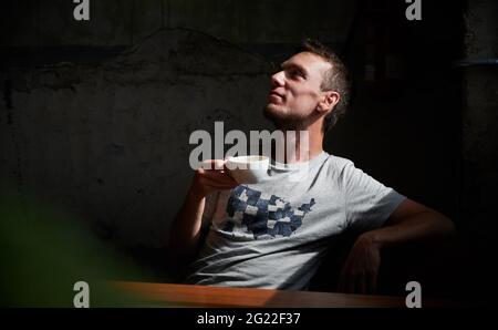Nahaufnahme eines hübschen jungen Mannes, der auf einem gemütlichen Stuhl sitzt und eine Tasse Kaffee hält und trinkt. Konzept der freien Zeit mit einem heißen Getränk zu genießen. Stockfoto