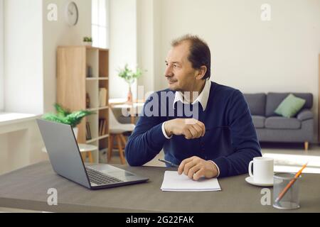 Nachdenklich lächelnder Erwachsener, der am Tisch sitzt und den Computer wegschaut Stockfoto