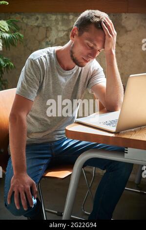 Junger frustrierter Mann in grauem T-Shirt, der auf einem Laptop sitzt und über die Arbeit nachdenkt. Konzept der Prozessarbeit im Laptop zu Hause oder im Café. Stockfoto