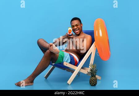 Verspielter schwarzer Mann, der sich mit einer Wasserpistole im Loungesessel entspannt und mit der Spielzeugpistole auf die Kamera über dem blauen Studiohintergrund zeigt Stockfoto