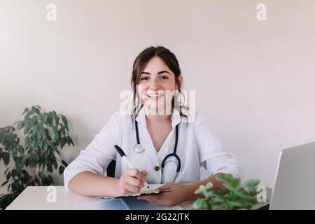 Lächelnde, hübsche Ärztin oder Krankenschwester in weißem Kleid mit Stethoskop am Arbeitsplatz, die Kamera anschaut und etwas in den Arzt des Patienten schreibt Stockfoto