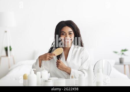 Tägliche Haarpflege zu Hause während des covid-19 Lockdown. Glückliche afroamerikanische Dame kämmt ihr lockiges Haar im Schlafzimmer Stockfoto