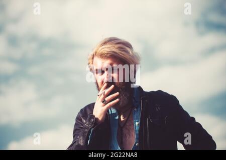 Trendiger, cooler junger Mann, der draußen steht und Zigarette raucht. Am blauen Himmel. Stockfoto