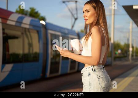 Junge Frau wartet auf den Stadtzug und telefoniert mit Kaffee in der Hand. Passagier an der Straßenbahnhaltestelle mit Smartphone. Konzept des öffentlichen Verkehrs und der Kommunikation. Hochwertige Fotos Stockfoto