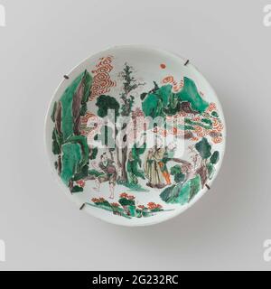 Saucer-dish with Four figures in a Rocky landscape. Scale of porcelain with round wall, painted in underglaze blue and on the glaze red, green, yellow, eggplant and black. On the front a continuous representation of four people, three in a group and one in the foreground, in a roty landscape with trees and clouds. Marked on the back with a shell in a double circle. With later iron suspension system. Six chips in the edge. Famille Verte. Stockfoto