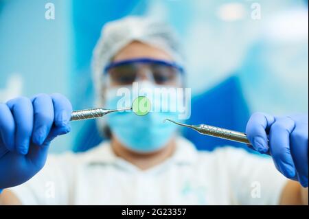 Zahnärztliche Instrumente in den Händen einer verschwommenen Zahnärztin, die auf einem verschwommenen blauen Hintergrund eine zahnärztliche Untersuchung macht Stockfoto