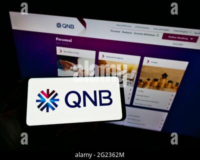 Person, die Mobiltelefon mit dem Logo der Finanzgesellschaft Qatar National Bank (QNB) auf dem Bildschirm vor der Business-Webseite hält. Konzentrieren Sie sich auf die Telefonanzeige. Stockfoto