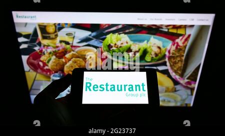 Person, die ein Mobiltelefon mit dem Logo des britischen Gastronomieunternehmens The Restaurant Group plc auf dem Bildschirm vor der Webseite hält. Konzentrieren Sie sich auf die Telefonanzeige. Stockfoto