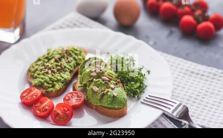 Veganes Avocado-Sandwich mit Tomaten und Microgreens auf einem weißen Teller auf grauem Hintergrund Stockfoto