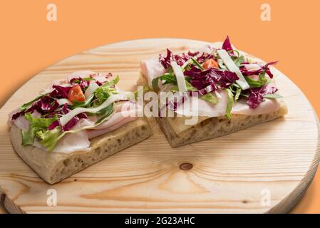 Italienische Focaccia mit Schinken, Salat, Käse und Tomaten. Zwei Scheiben auf Holzschneidebrett isoliert auf orangefarbenem Hintergrund Stockfoto