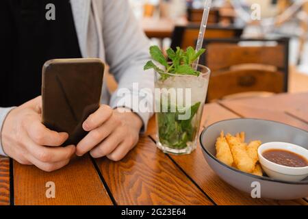 Nahaufnahme Foto von Menschenhänden am Couchtisch mit Smartphone neben kühlenden Mojito-Cocktails, mobiles Gerätekonzept Stockfoto