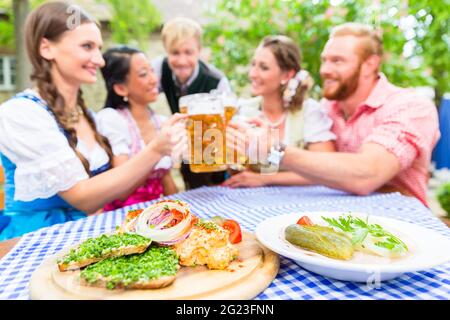 Fünf Freunde, Frauen und Männer, sitzen im Biergarten klirren Gläser, die Bayerischen Vorspeisen auf dem Tisch Stockfoto