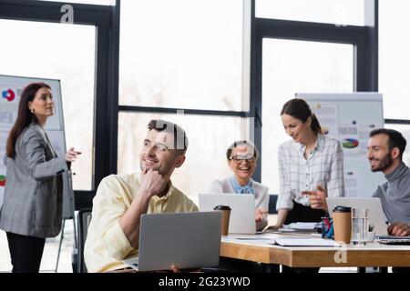 Lächelnder Geschäftsmann mit Laptop in der Nähe verschwommener multiethnischer Kollegen Stockfoto