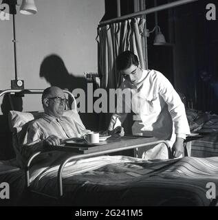 Historischer, junger Arzt aus den 1960er Jahren, der abends in einer Stationsschicht arbeitet, um einem älteren männlichen Patienten zu helfen, sich in seinem Bett wohler zu fühlen, indem er den Rahmen des Metallbetthalters für sein Esstablett justiert, Schottland, Großbritannien. Stockfoto