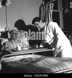 Historischer, junger Arzt aus den 1960er Jahren, der abends in einer Stationsschicht arbeitet, um einem älteren männlichen Patienten zu helfen, sich in seinem Bett wohler zu fühlen, indem er den Rahmen des Metallbetthalters für sein Esstablett justiert, Schottland, Großbritannien. Stockfoto