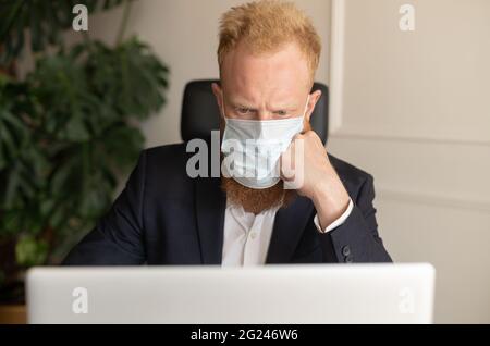Nachdenklicher rothaariger Geschäftsmann in schickem Freizeitanzug, der eine schützende Gesichtsmaske trägt und einen Laptop-Computer im modernen Büro verwendet, der sich um die Finanzkrise aufgrund einer Pandemie sorgt Stockfoto
