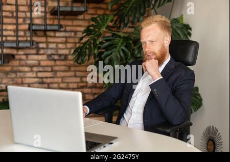 Aufmerksamer rothaariger Geschäftsmann in elegantem Freizeitanzug, der auf den Laptop-Bildschirm starrt, über Strategie nachdenkt, schwierige Aufgaben löst, nachdenklicher Mann, der im modernen Büro einen Laptop benutzt Stockfoto