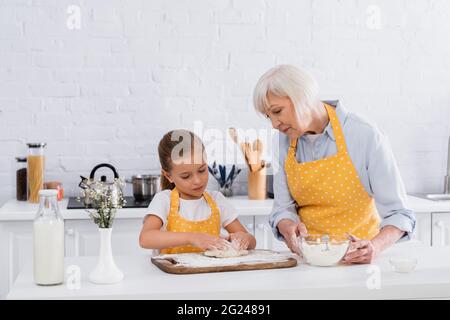 Oma hält Mehl, während das Kind Teig in der Nähe von Milch in der Küche macht Stockfoto