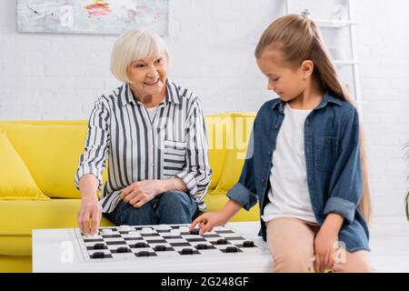 Lächelnde ältere Frau spielt Dame Spiel mit verschwommenen Enkelin Stockfoto