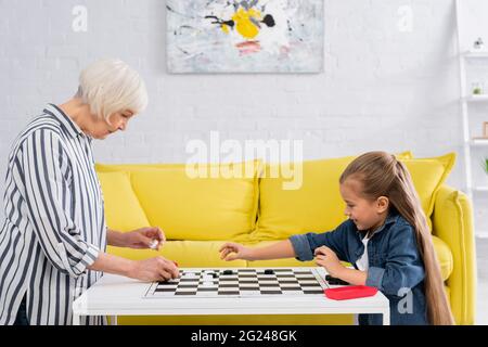 Seitenansicht einer älteren Frau, die mit der Enkelin im Wohnzimmer Dame spielte Stockfoto