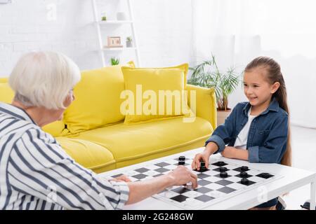 Lächelndes Kind spielt Dame mit verschwommener Großmutter zu Hause Stockfoto