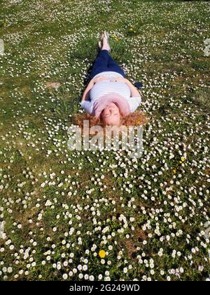 Schöne rothaarige reife Frau in ihren Fünfzigern liegt zufrieden im Gras und genießt die Sonne und fühlt sich in einem Feld von Gänseblümchen gut. Stockfoto