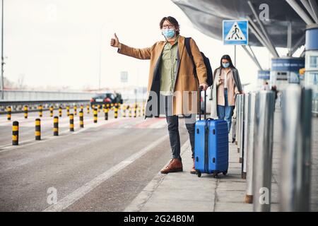 Stilvoller junger Mann in medizinischer Maske, der am Flughafen ein Taxi nimmt Stockfoto