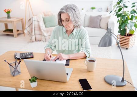 Foto einer ruhigen, fokussierten, gealterten Frau, die hinter dem Schreibtisch sitzt, geballter Notizblock, der sich Notizen von zu Hause aus machen kann Stockfoto