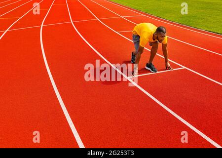 Seitenansicht des aktiven, konzentrierten jungen, afroamerikanischen Sprinters in gelber Sportkleidung, der zum Laufen auf der Sportstrecke bereit ist. Läufer Stockfoto