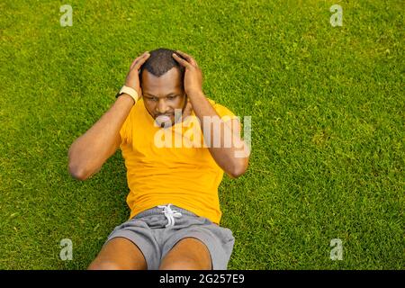 Junger sportlich fokussierter afroamerikanischer Mann, der auf grünem Gras liegt und morgens draußen trainiert, gesunder positiver aktiver Schwarzer, der ABS trainiert Stockfoto