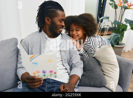 Vater-Tochter-Beziehung. Papa freut sich, eine Grußkarte mit einer Kinderzeichnung von seiner Tochter bis zum Vatertag zu erhalten, die auf einem Sofa am Morgen sitzt Stockfoto