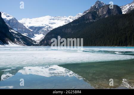 Wunderschöne Szene mit Lake Louise, teilweise gefroren und die Berge um sie herum spiegeln sich darin. Sonniger Tag im Frühling. Banff National Park, Alberta, Kanada Stockfoto