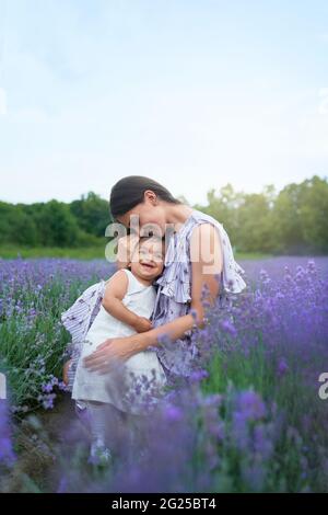 Seitenansicht der jungen Mutter, die Kleid trägt, das zwischen schönen violetten Blumen sitzt. Frau posiert mit kleinen lächelnden Baby-Tochter auf den Knien in Lavendelfeld. Konzept der Natur, Mutterschaft. Stockfoto