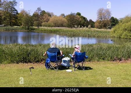 Ein Paar sitzt auf Stühlen und picknickt an einem See, Blick nach hinten, an einem sonnigen Frühlingstag, Cambridgeshire England Stockfoto