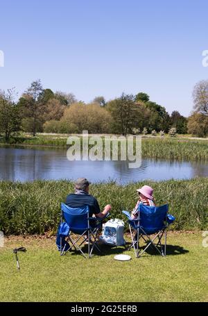 Ein Paar sitzt auf Stühlen und picknickt an einem See, Blick nach hinten, an einem sonnigen Frühlingstag, Cambridgeshire England Stockfoto