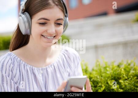 Lächelnde Junge Studentin Auf Dem College Campus Streamt Musik Vom Mobiltelefon Auf Kabellose Kopfhörer Stockfoto